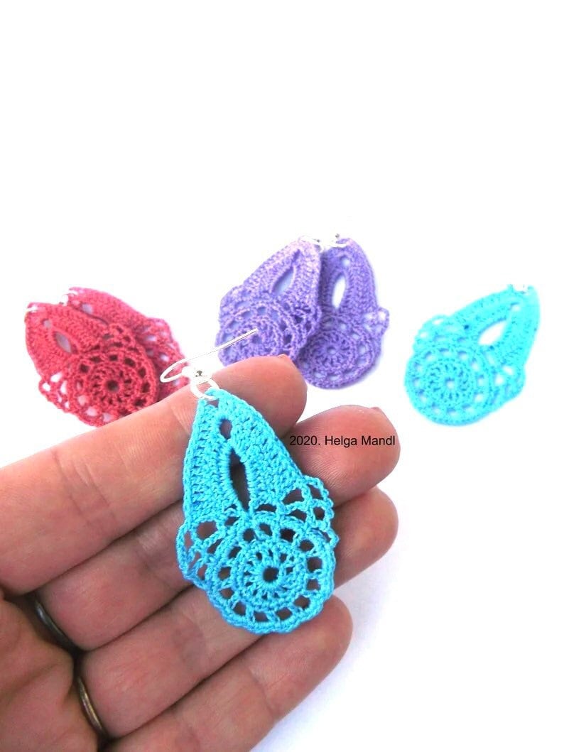 Puede incluir: Primer plano de un pendiente de ganchillo azul claro sostenido en una mano, con otros pendientes de ganchillo en tonos rojos, morados y turquesas en el fondo. Los pendientes tienen un dise&ntilde;o calado delicado. La imagen est&aacute; sobre un fondo blanco.