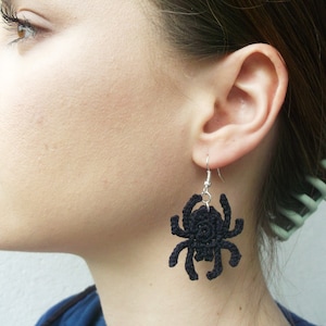 May include: Black crocheted spider earrings hanging from silver hooks. The earrings are worn by a person with brown hair.