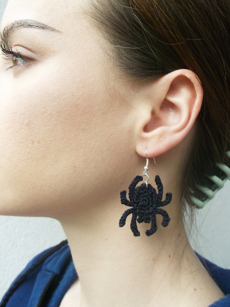 May include: Black crocheted spider earrings hanging from silver hooks. The earrings are worn by a person with brown hair.
