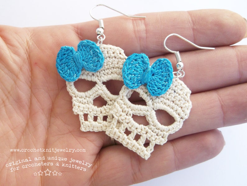 May include: A pair of crocheted skull earrings with blue bows. The earrings are white and have a delicate, lacy design. The bows are made of a bright blue yarn.