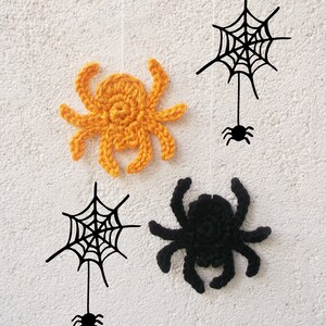 May include: Four Halloween decorations hanging from strings against a white wall. Two black crocheted spiders and two black web designs with a small black spider at the bottom. One spider is orange.