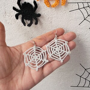 May include: A pair of white crocheted spiderweb earrings with silver hooks. The earrings are hanging from a hand.
