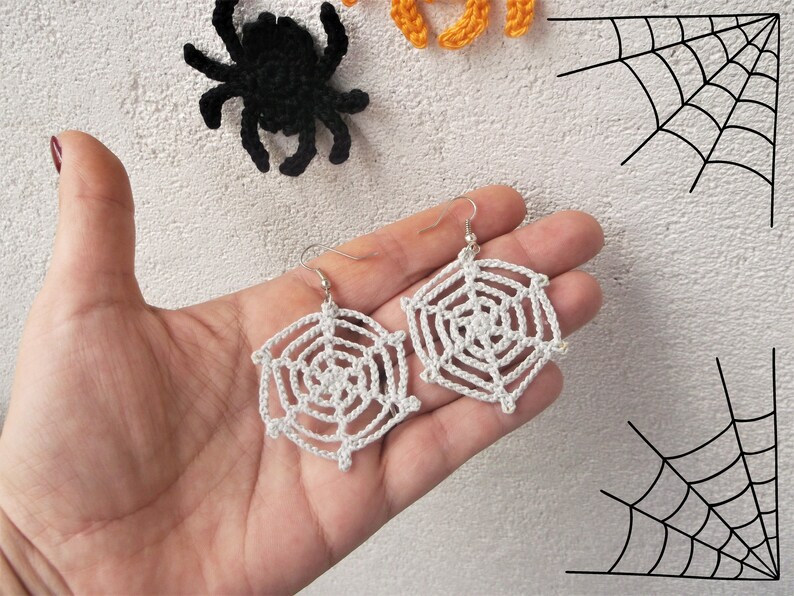 May include: A pair of white crocheted spiderweb earrings with silver hooks. The earrings are hanging from a hand.
