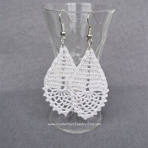 May include: White crochet teardrop earrings with silver hooks. The earrings are hanging from a glass stand.