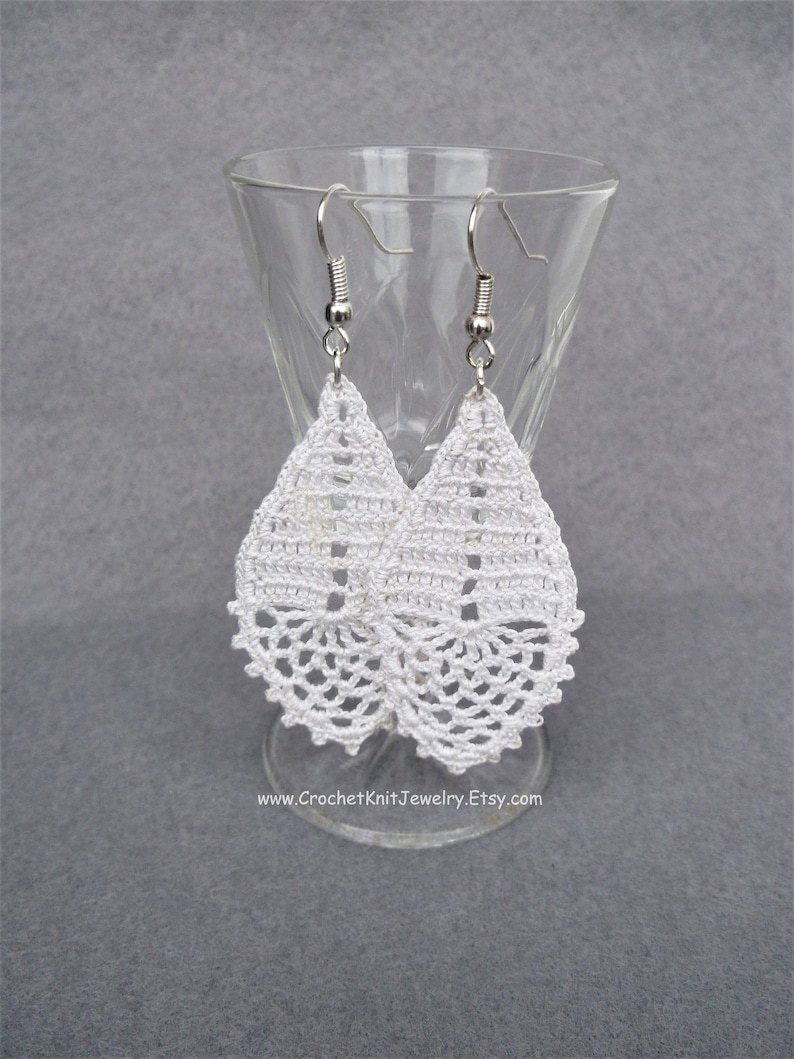 May include: White crochet teardrop earrings with silver hooks. The earrings are hanging from a glass stand.