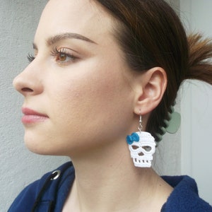 May include: White crocheted skull earrings with a blue bow. The earrings are hanging from a person's ear.