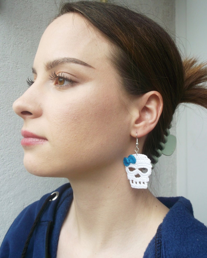 May include: White crocheted skull earrings with a blue bow. The earrings are hanging from a person's ear.