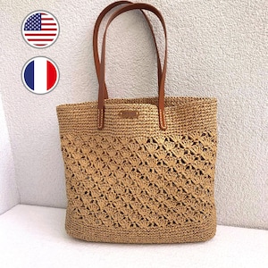 May include: A woven straw tote bag with brown leather handles. The bag features a decorative openwork pattern on the lower half and a small "handmade" tag. The bag is a natural tan color.