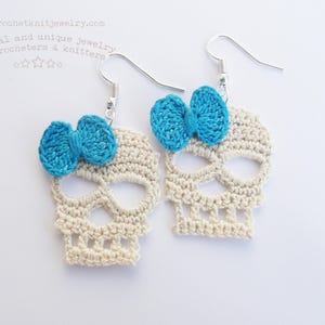 May include: A pair of crocheted skull earrings with blue bows. The earrings are made of white yarn and have a delicate, intricate design.