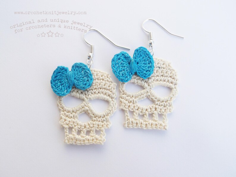 May include: A pair of crocheted skull earrings with blue bows. The earrings are made of white yarn and have a delicate, intricate design.