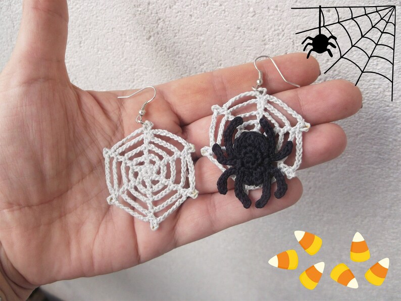 May include: A pair of crocheted spider web earrings with a black crocheted spider on one earring. The earrings are held in a hand against a white background.