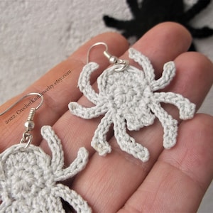 May include: A pair of crocheted spider earrings with silver hooks. The spiders are white and have eight legs.