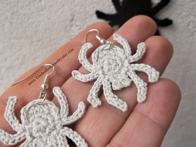 May include: A pair of crocheted spider earrings with silver hooks. The spiders are white and have eight legs.