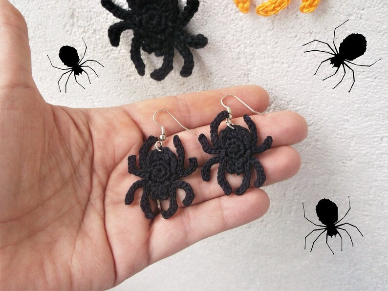 May include: A pair of black crocheted spider earrings hanging from silver hooks. The earrings are held in a hand against a white background.