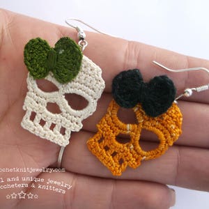 May include: A pair of crocheted skull earrings. One earring is white with a green bow and the other is orange with a black bow. Both earrings have silver hooks.