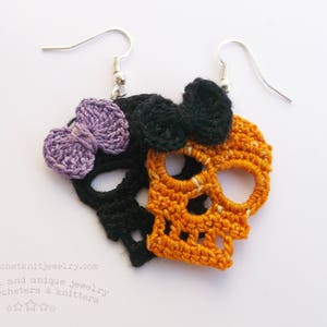 May include: Crocheted skull earrings with a black skull and a purple bow on one side and an orange skull on the other side. The earrings are attached to silver hooks.