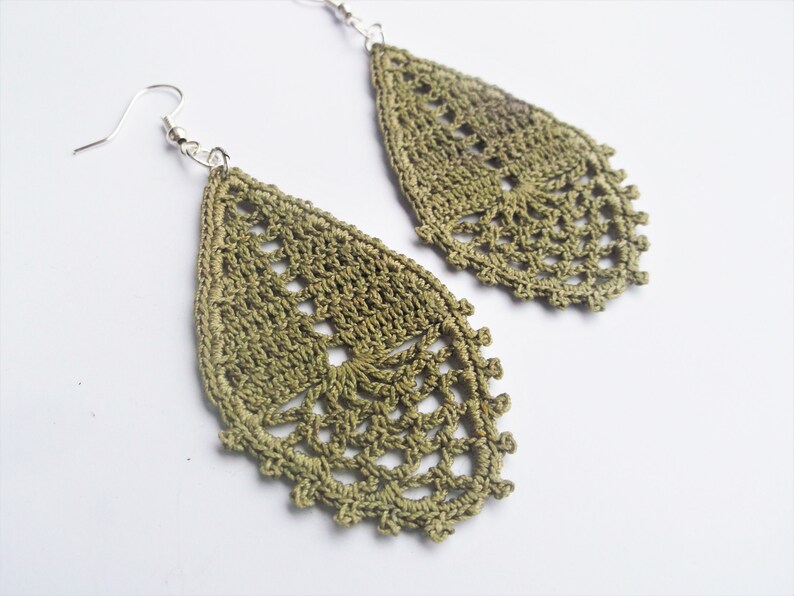 May include: A pair of green crochet earrings with a teardrop shape. The earrings are made with a delicate, lacy pattern.
