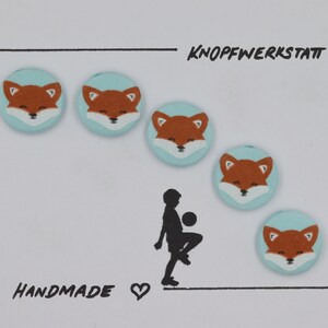 May include: Five handmade buttons with a light blue background and a fox face design. The fox faces are orange with white accents. The buttons are arranged in a semi-circle with the text "KNOPFWERKSTATT" and "HANDMADE" visible.