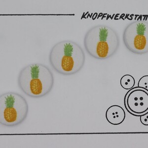 Könnte beinhalten: Vier weiße Stoffknöpfe mit Ananas-Motiv. Die Ananas sind gelb mit grünen Spitzen. Die Knöpfe sind diagonal auf weißem Hintergrund angeordnet. Der Text "KNOPFWERKSTATT" ist oben im Bild mit schwarzem Stift geschrieben.