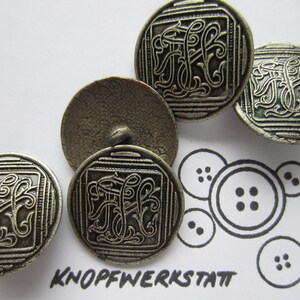 Könnte beinhalten: Vier silberfarbene Knöpfe mit einem verzierten Design, das einen stilisierten Buchstaben "F" in der Mitte zeigt. Die Knöpfe befinden sich auf einer weißen Oberfläche mit einer Zeichnung von Knöpfen.