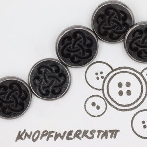 Peut inclure: Cinq boutons argentés avec des motifs de nœuds celtiques noirs. Les boutons sont disposés en rangée, le plus grand en haut et le plus petit en bas. Les boutons sont sur un fond blanc. Le texte "KNOFFWERKSTATT" est écrit en noir sous les boutons.