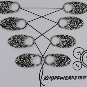 Puede incluir: Ocho topes de botón de metal plateados con un diseño floral. Los topes están dispuestos en dos filas de cuatro, con la fila superior ligeramente desplazada de la fila inferior. Los topes están sobre un fondo blanco con el texto "KNOFFWERKSTATT" impreso en negro en la parte inferior.
