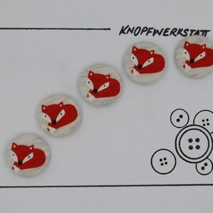 May include: Five red and white fabric buttons with a fox design. The buttons are arranged in a line on a white background. The text "KNOFFWERKSTATT" is written in black ink above the buttons.