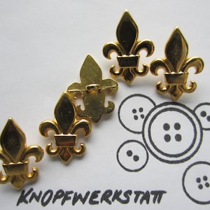Könnte beinhalten: Fünf goldfarbene Metallknöpfe in Form einer Fleur-de-lis. Die Knöpfe sind in einem Halbkreis auf einer weißen Oberfläche angeordnet. Die Knöpfe sind alle gleich groß und gleich geformt.