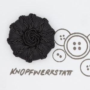 Pode incluir: Um botão coberto de tecido preto com um desenho de rosa no centro. O botão é rodeado por um padrão decorativo de corda. O texto "KNOPFWERKSTATT" está escrito abaixo do botão.