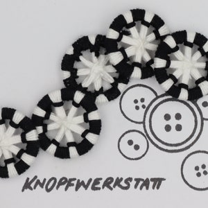 May include: Five black and white striped fabric buttons with a white center and a starburst design. The buttons are arranged in a curved line. The word "KNOFFWERKSTATT" is written in black below the buttons.