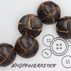 Könnte beinhalten: Sechs dunkelbraune, kugelförmige Knöpfe mit einem strukturierten, genähten Design. Sie liegen auf einer weißen Oberfläche. Das Bild enthält eine Grafik von Knopflinien und das Wort "KNOPFWERKSTATT".