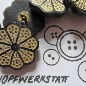Könnte beinhalten: Eine Sammlung von sechs dekorativen Knöpfen mit einem floralen Design. Die Knöpfe sind aus Holz und haben eine schwarze und goldene Oberfläche. Die Knöpfe sind in einem Kreis um das Wort "Knopfwerkstatt" angeordnet.