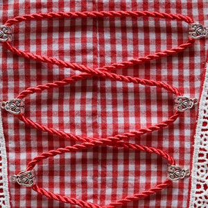 Puede incluir: Primer plano de una tela a cuadros rojos y blancos con detalles de cuerda roja. La cuerda se sujeta con cierres metálicos plateados. Ribete de encaje blanco con motivos florales bordea la tela.