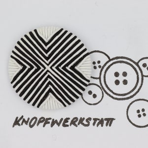 Pode incluir: Um botão redondo com listras pretas e brancas com um design tecido. O botão é redondo e tem um centro elevado. O botão é rodeado por um fundo branco com o texto "KNOPFWERKSTATT" escrito em preto.