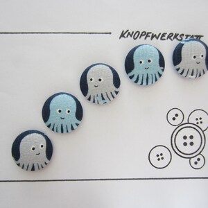 May include: Five blue and white fabric buttons with an octopus design. The buttons are arranged in a row on a white background. The text "KNOFFWERKSTAT" is written in black ink above the buttons.