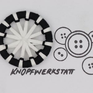 Op de afbeelding: Een zwarte en witte knoop met een geweven ontwerp. De knoop is rond en heeft een wit midden met zwarte spaken die vanuit het midden stralen. De knoop is omgeven door een zwart-wit gestreepte rand. Het woord "KNOFFWERKSTATT" staat in het zwart onder de knoop.