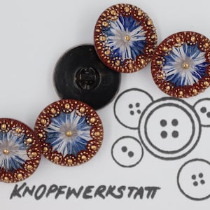 Può includere: Quattro bottoni decorativi con un motivo floreale blu e bianco, circondati da un bordo dorato. I bottoni sono su una superficie bianca con il testo "KNOPFWERKSTATT" scritto in nero.