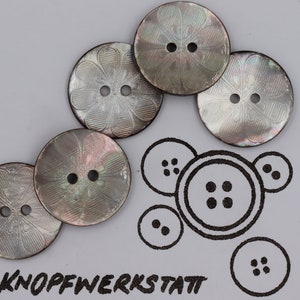 Puede incluir: Cinco botones iridiscentes de nácar con un diseño floral. Los botones son redondos y tienen dos agujeros cada uno. Los botones están dispuestos en un semicírculo.