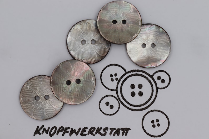 May include: Five iridescent mother-of-pearl buttons with a floral design. The buttons are round and have two holes each. The buttons are arranged in a semi-circle.