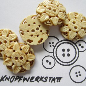 Könnte beinhalten: Fünf cremefarbene, kunstvoll geschnitzte Knöpfe mit Blumenmuster. Die Knöpfe sind auf einer weißen Oberfläche mit einer schwarz-weißen Zeichnung von Knöpfen angeordnet.