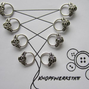 Puede incluir: Ocho marcadores de punto de plata en forma de rosa con bucles. Los marcadores están dispuestos en un patrón sobre un fondo blanco. La palabra "KNOPFWERKSTATT" está escrita en tinta negra sobre el fondo.