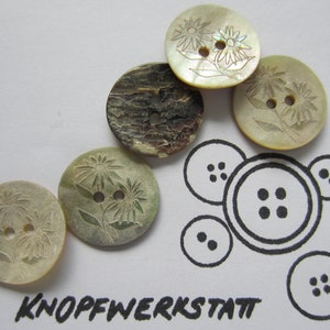 Puede incluir: Una colección de botones redondos en varios colores y texturas. Algunos botones son de color claro con diseños florales grabados, mientras que otros tienen una apariencia más oscura y texturizada. El texto "KNOPFWERKSTATT" está impreso en negro en la parte inferior.