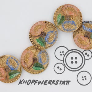 May include: Five round, gold-rimmed buttons with a floral design. The buttons have a pink, blue, and green floral pattern on a white background. The buttons are arranged in a curved line.