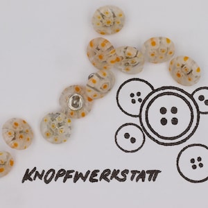 May include: A collection of clear, oval-shaped buttons with yellow and white floral patterns. Some buttons have a metal shank. The image also includes a graphic of several button designs and the text "KNOPFWERKSTATT".