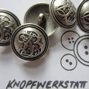 Pode incluir: Cinco botões de metal prateado com um design decorativo que apresenta uma letra B estilizada. Os botões têm um acabamento liso e brilhante.