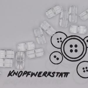 May include: Clear, rectangular beads and a drawing of various button designs are scattered on a light gray surface. The text "KNOPFWERKSTATT" is printed in black below the beads.
