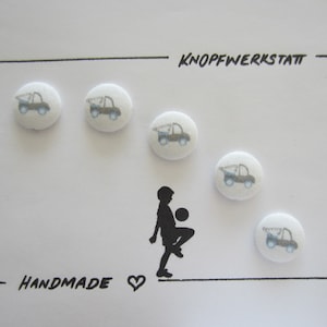 May include: Handmade buttons with a blue truck design on a white background. The buttons are arranged in a line, with a silhouette of a person kicking a ball in the middle. The text "KNOPFWERKSTATT" and "HANDMADE" are written on the card.