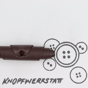 May include: A brown leather strip rolled up into a coil. The leather has a textured surface. The coil is on a white surface with a drawing of buttons and the text "KNOFFWERKSTATT".