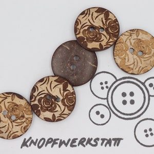 Könnte beinhalten: Fünf braune Kokosnuss-Knöpfe mit floralem Muster. Die Knöpfe haben jeweils zwei Löcher. Die Knöpfe sind in einem Halbkreis angeordnet. Das Wort "KNOFFWERKSTATT" ist in schwarzer Tinte am unteren Rand des Bildes geschrieben.