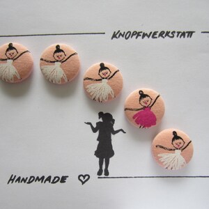 Pode incluir: Seis botões de tecido feitos à mão com fundo rosa e design de bailarina. Os botões estão dispostos em semicírculo com o texto "KNOPFWERKSTATT" e "HANDMADE" na parte superior e inferior da imagem.